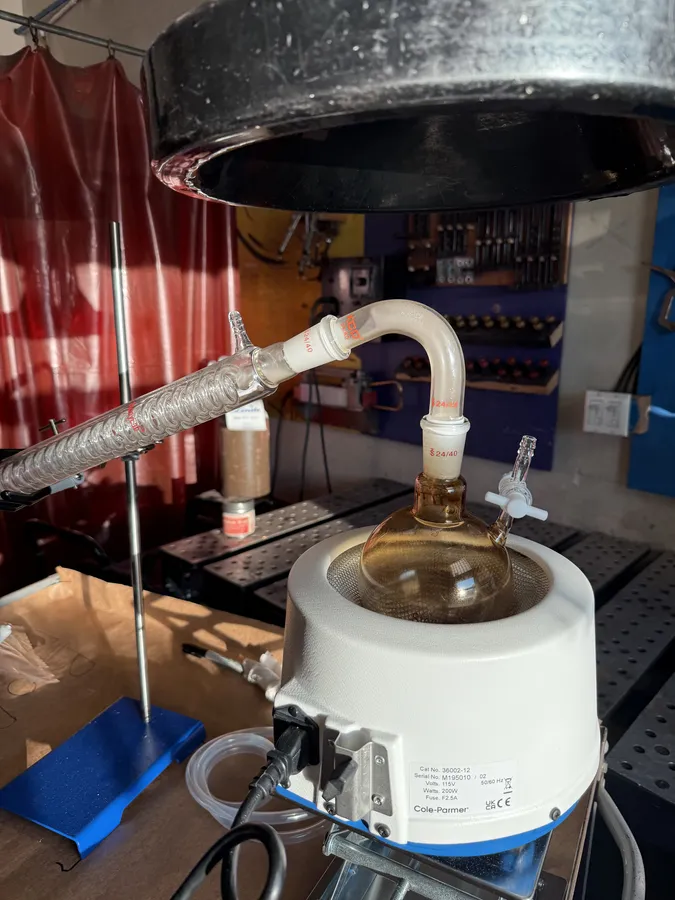 The flask on the heating mantle under a fume hood, brown liquid inside, the condenser leading away