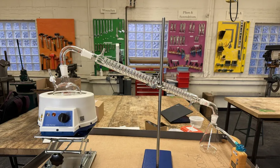 Bench pyrolysis setup on a shop bench: heating mantle and flask on a lab jack, a long glass condenser on a ring stand, and a receiving flask